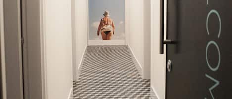 A hallway with a geometric patterned floor, a door with the word "WELCOME" on it, and a view of a beach through a window at the end of the hallway.