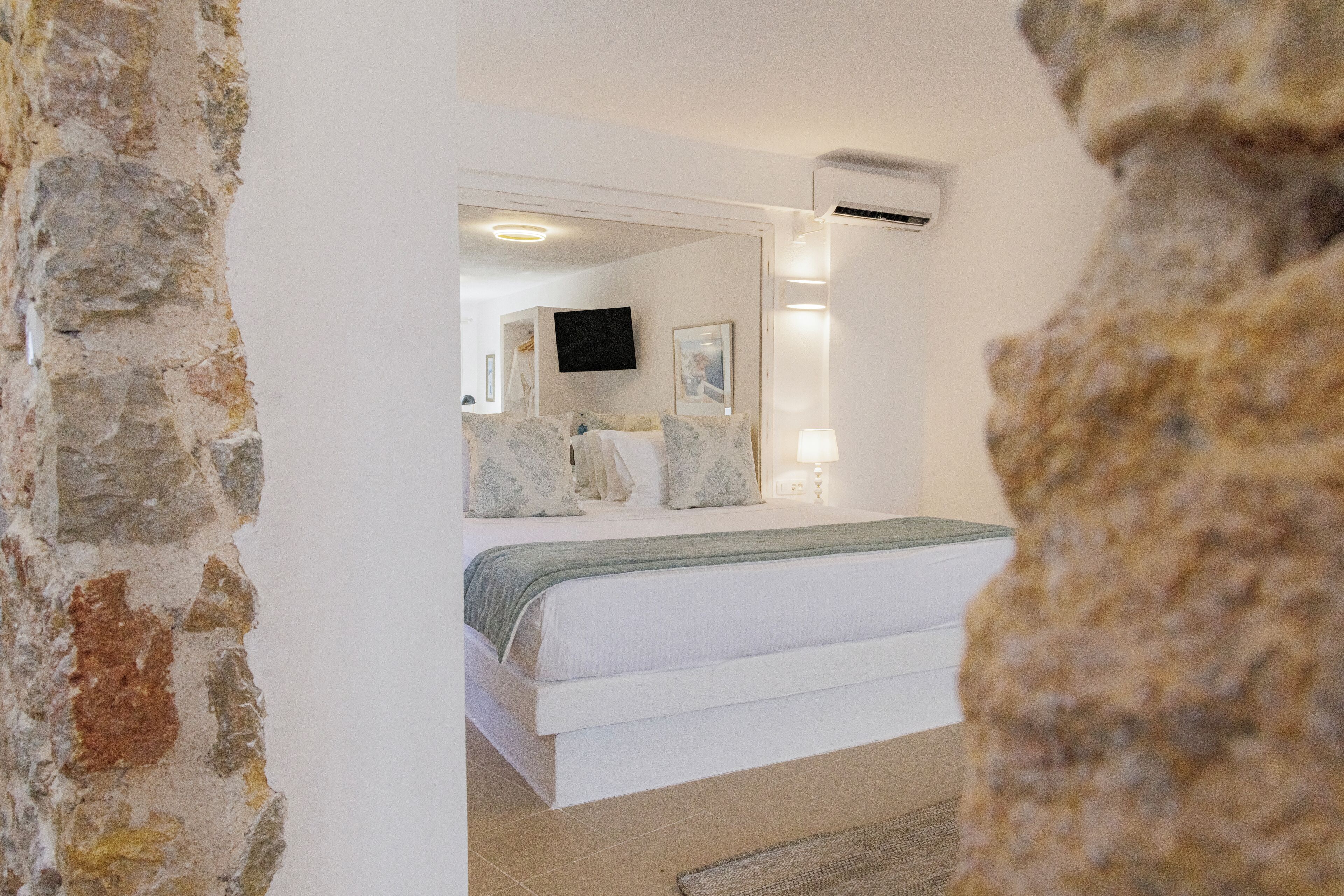 A bedroom with a bed, a flat-screen TV, wall-mounted lights, and a stone wall.