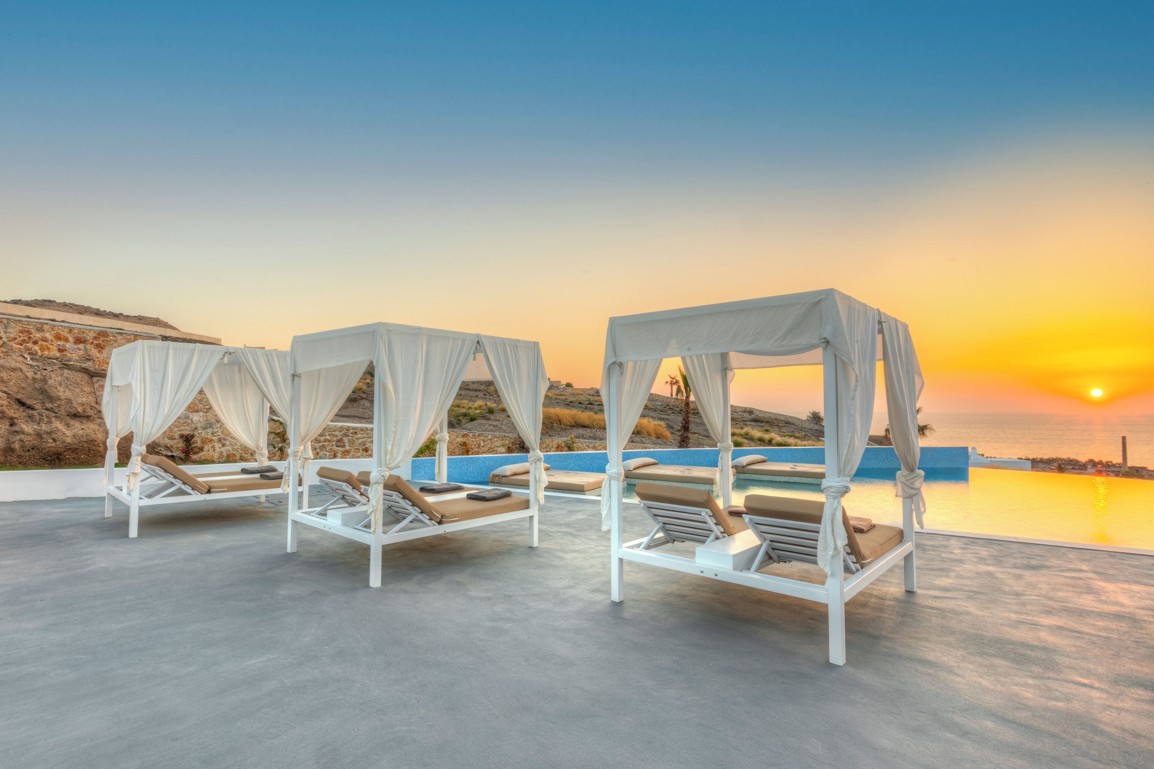 A rooftop terrace with four white canopy beds, a pool, and a sunset view.