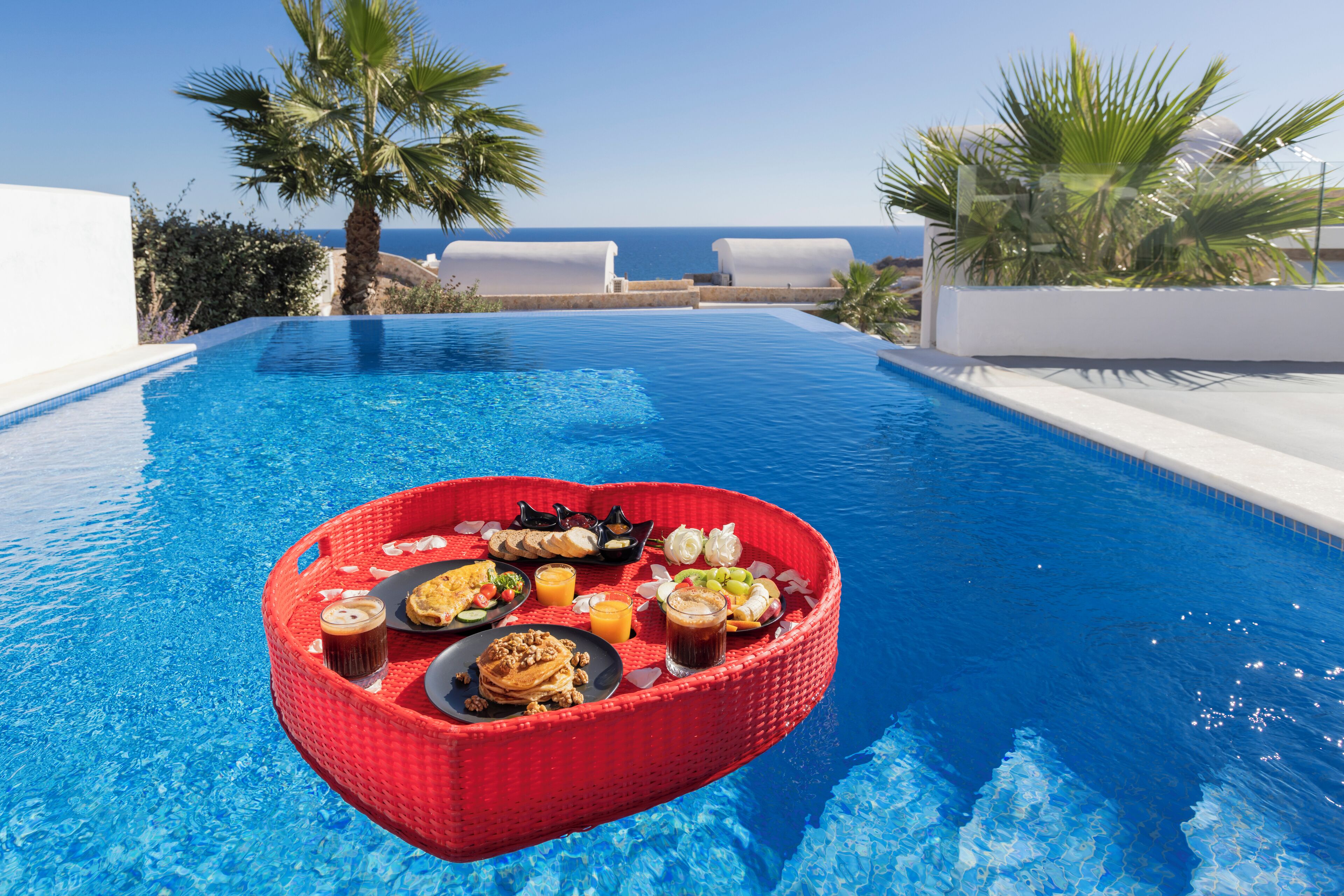 A poolside breakfast setup with a heart-shaped table, a variety of food and drinks, and a view of the sea.