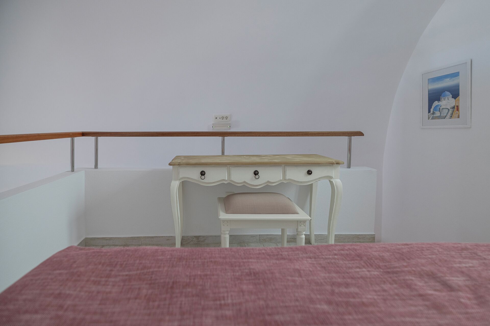 A bedroom with a bed, a white desk with a chair, a wooden railing, and a framed picture on the wall.