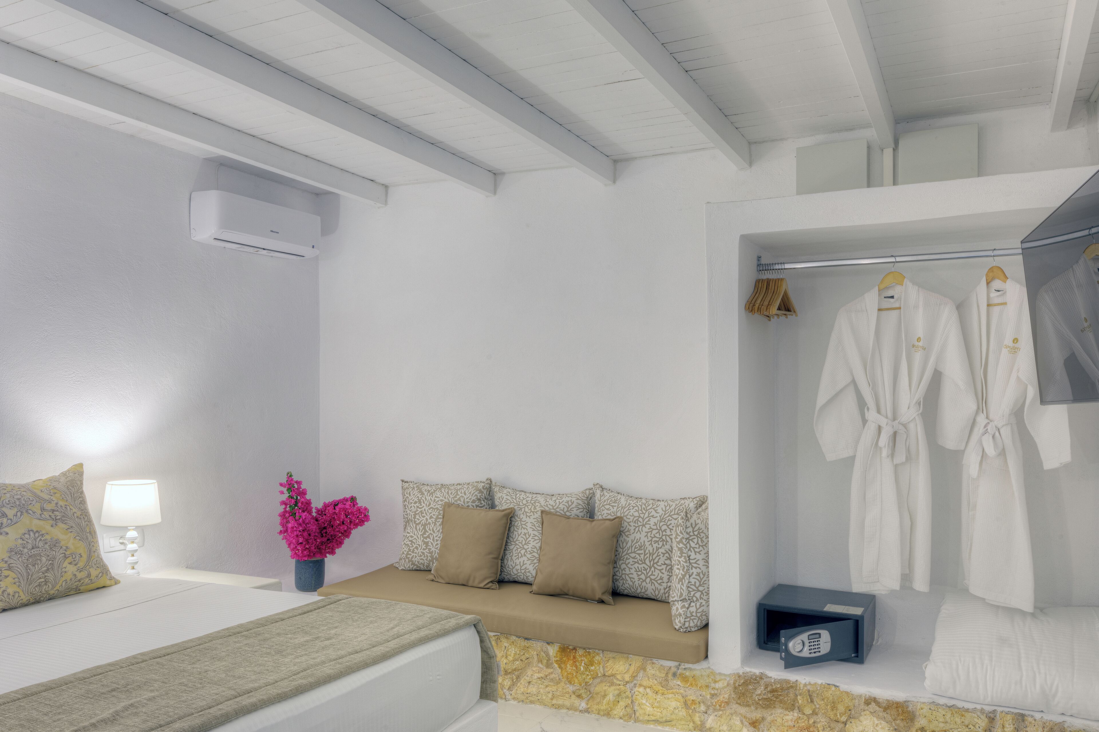 A modern hotel room with a bed, a bench with pillows, a vanity with a mirror, and a robe rack with robes.
