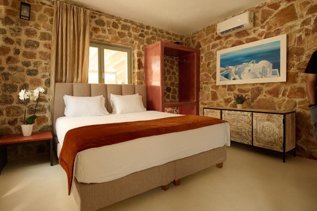 A bedroom with a stone wall, a bed with white bedding, a red bench, a wooden cabinet, and a painting.