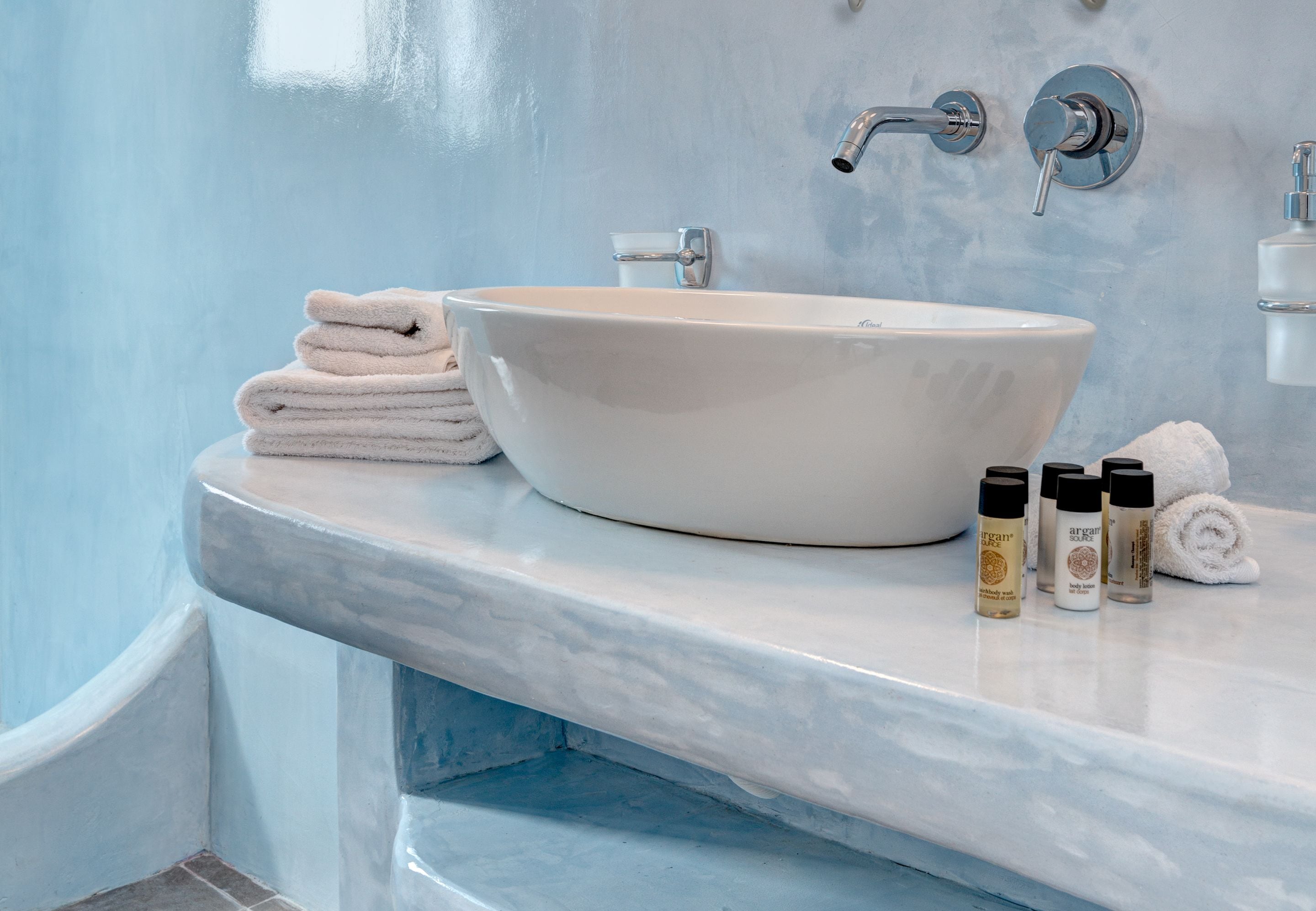 A bathroom with a white ceramic sink, two faucets, a towel stack, and three bottles of lotion on the countertop.