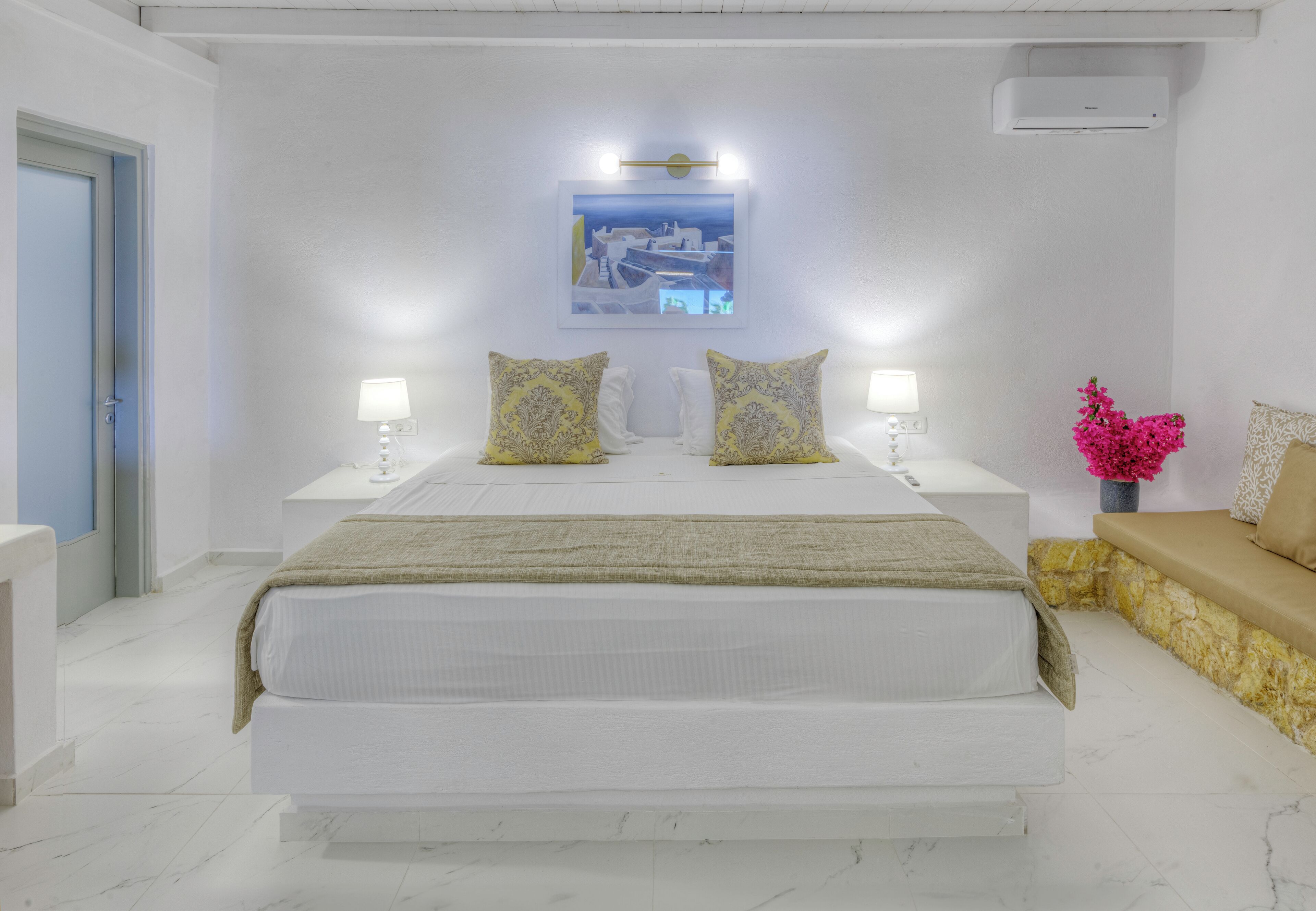 Una camera d'albergo con un letto grande, due abat-jour, un condizionatore d'aria a parete e un tavolino con un vaso di fiori.