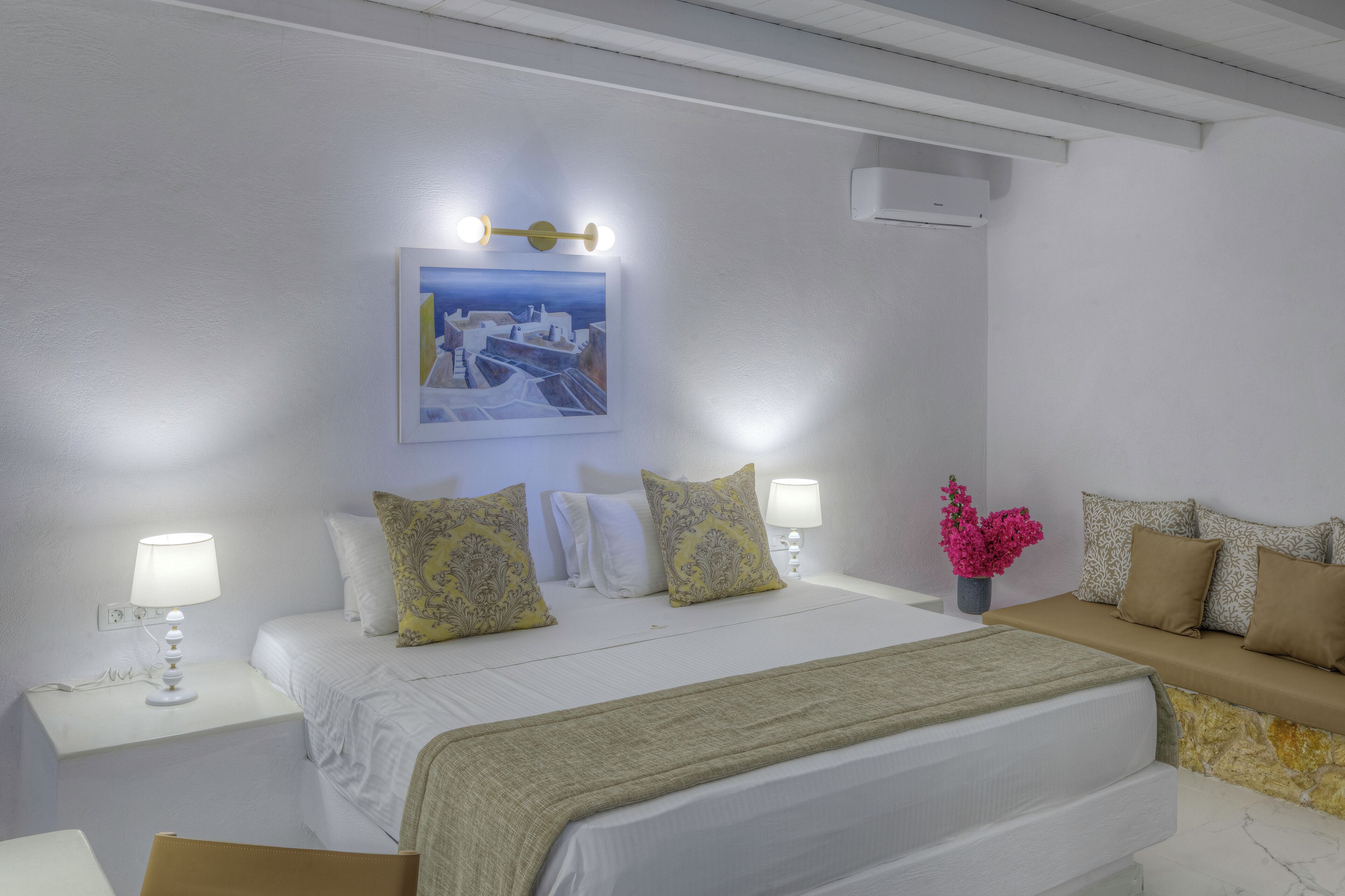 Ein Hotelzimmer mit einem Bett, Nachttischen, einer Bank, einer Wand-Klimaanlage und einem gerahmten Bild über dem Bett.