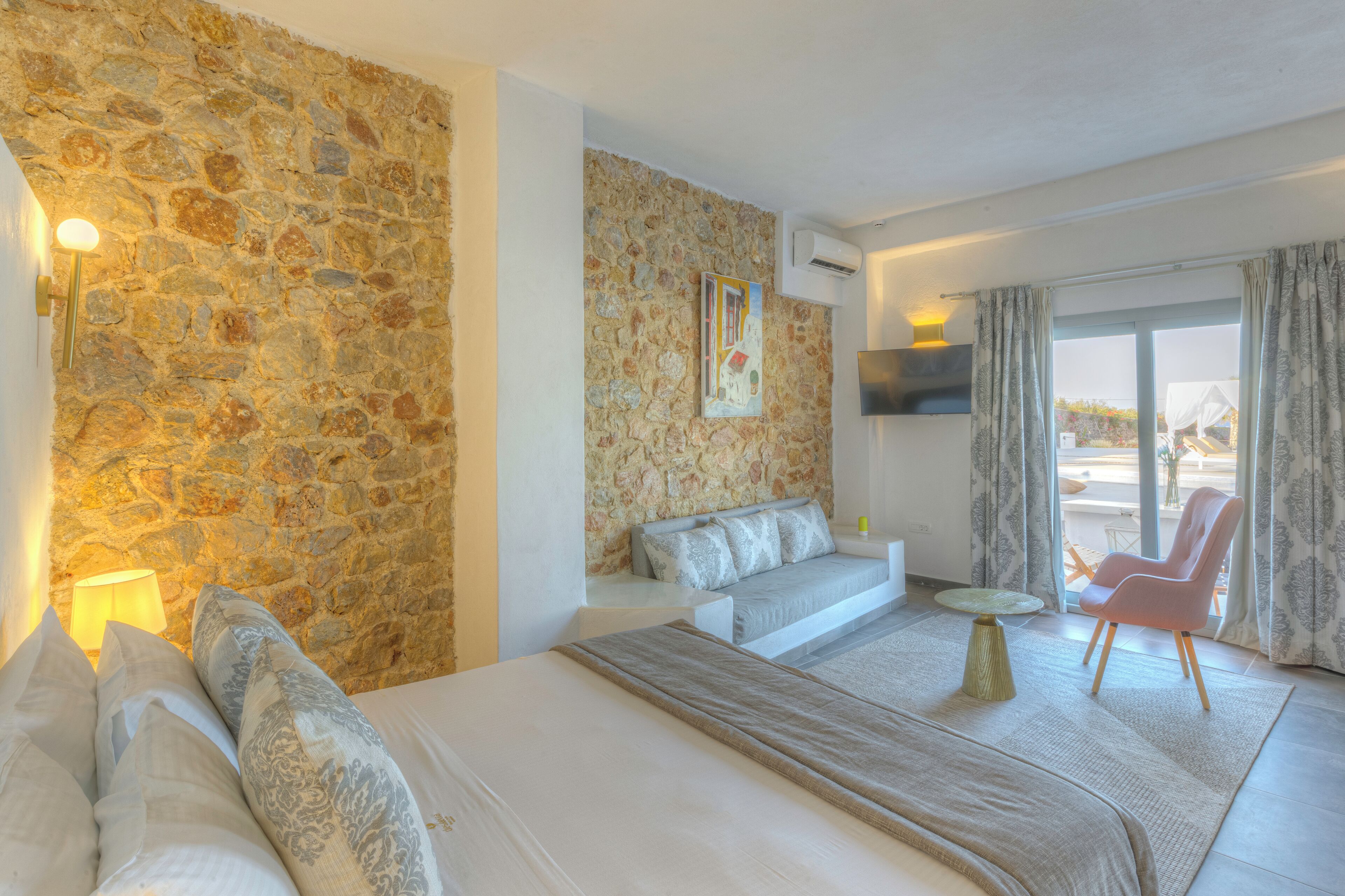 A modern hotel room with a stone wall, a sofa, a bed, a small round table, and a pink chair.