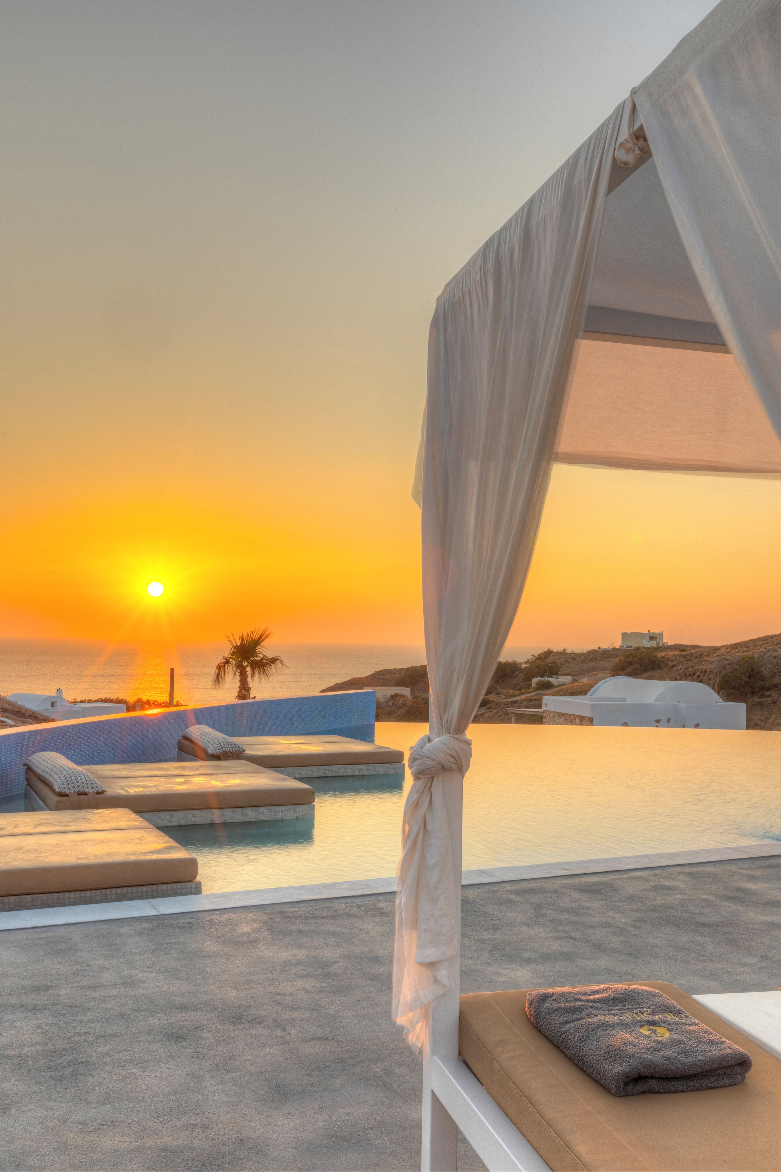A sunset view from a balcony with a pool, white curtains, and a towel on a bench.