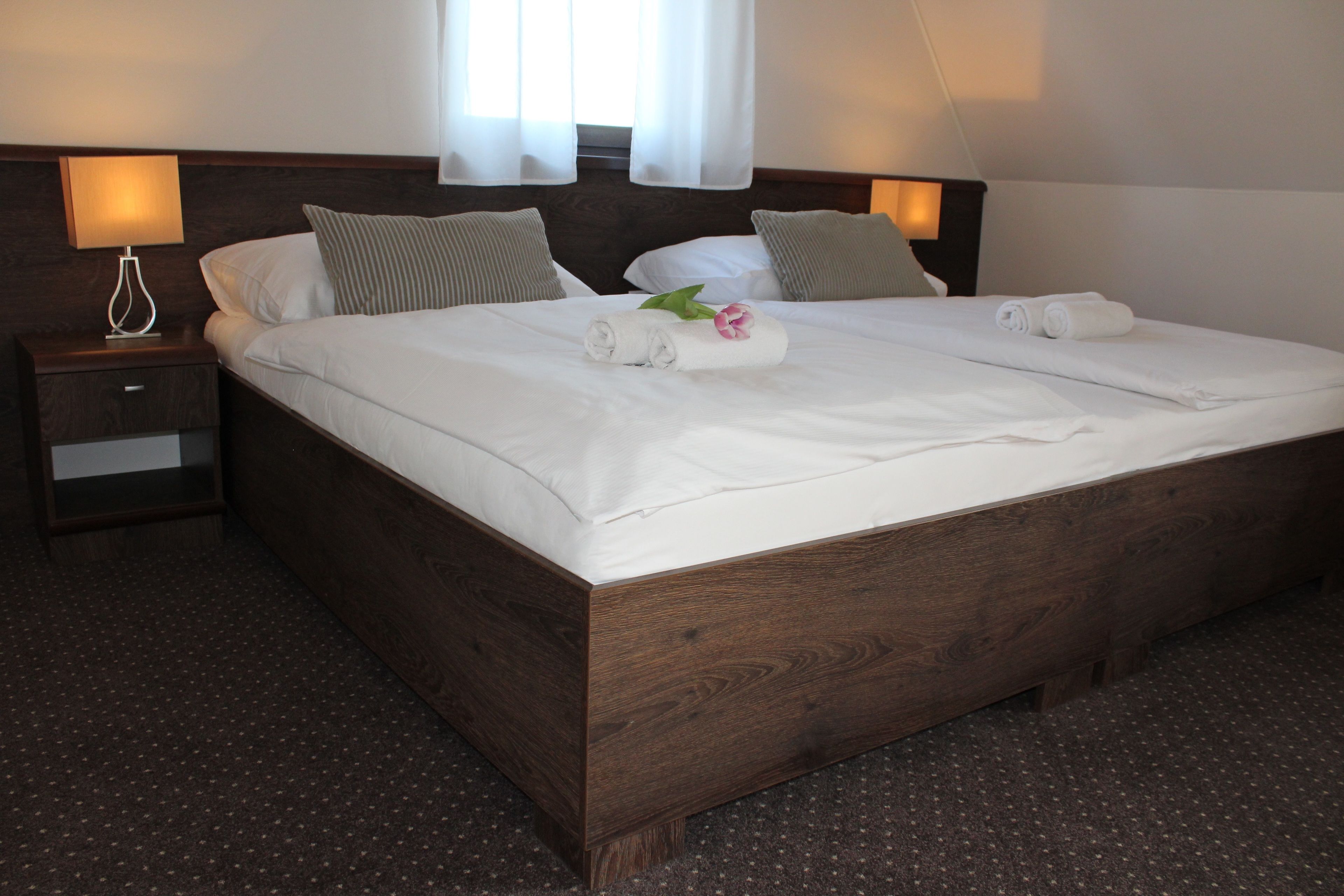 A neatly made double bed with white linens, two pillows, and a rolled towel with a flower. There is a wooden nightstand with a lamp beside the bed.