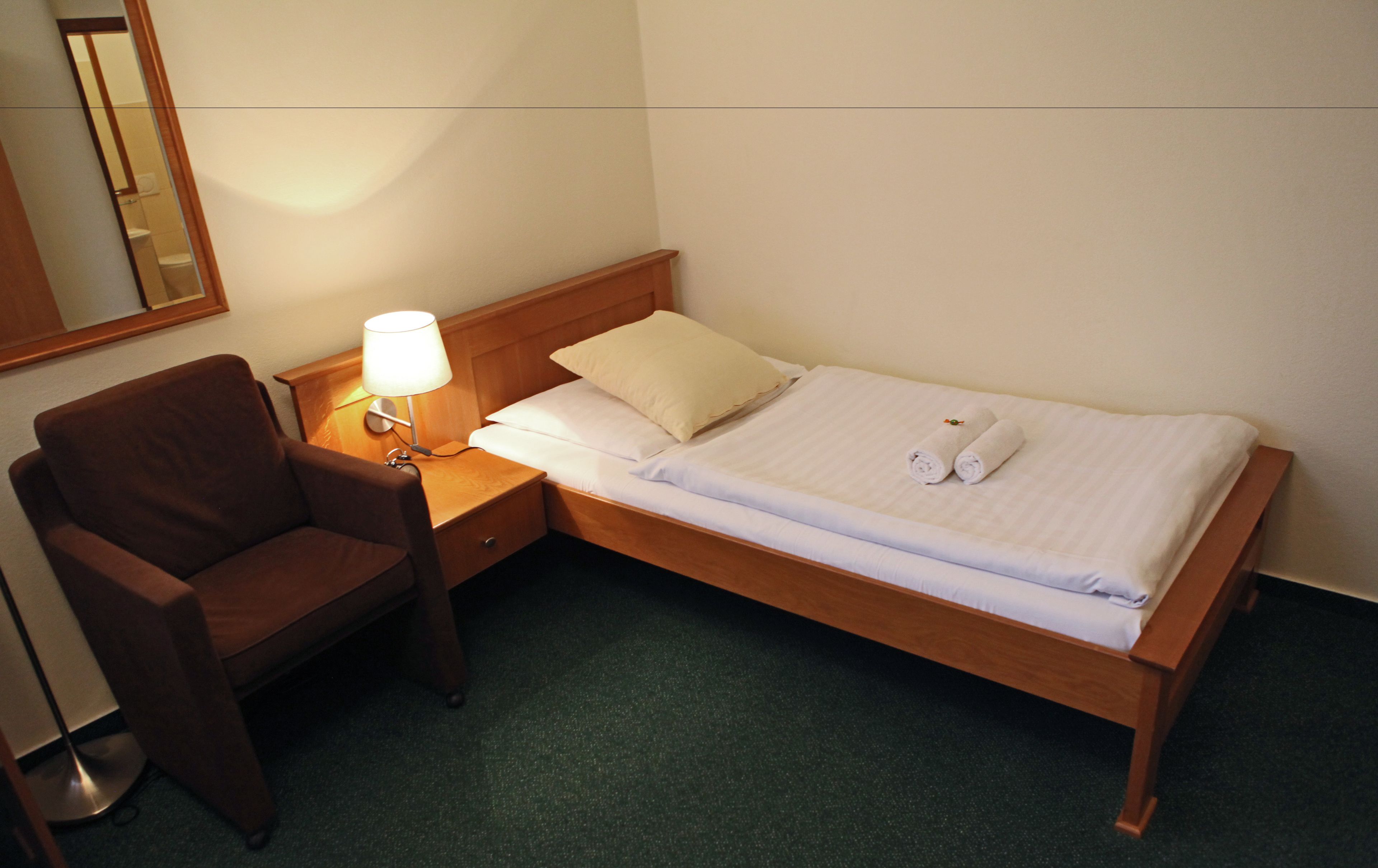 A single bed with white linens, a wooden headboard, a bedside table with a lamp, and a brown armchair.