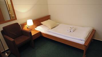 A single bed with white linens, a wooden headboard, a bedside table with a lamp, and a brown armchair.