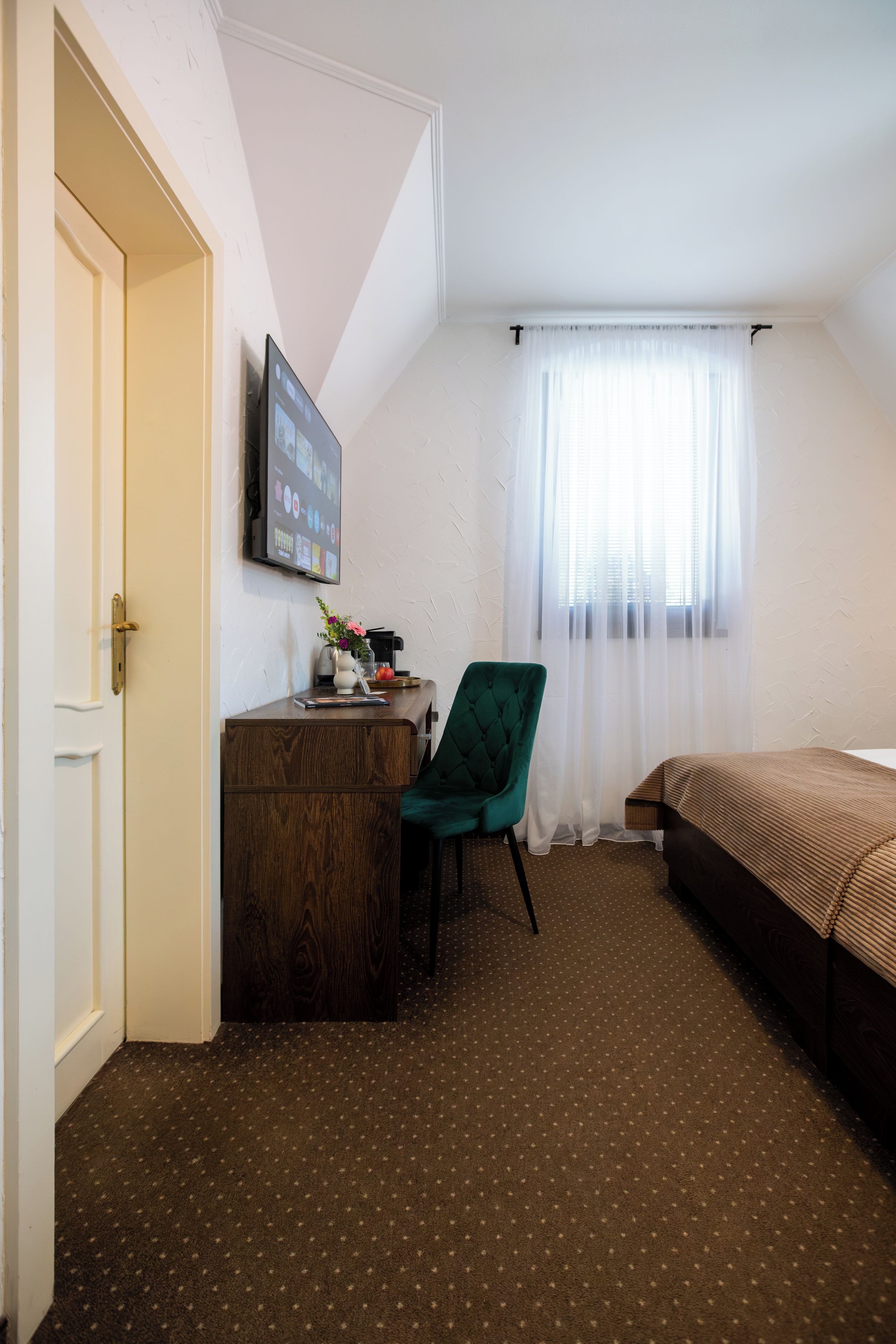A hotel room with a wooden desk, a green chair, a bed, and a window with sheer curtains.