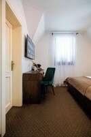 A hotel room with a wooden desk, a green chair, a bed, and a window with sheer curtains.
