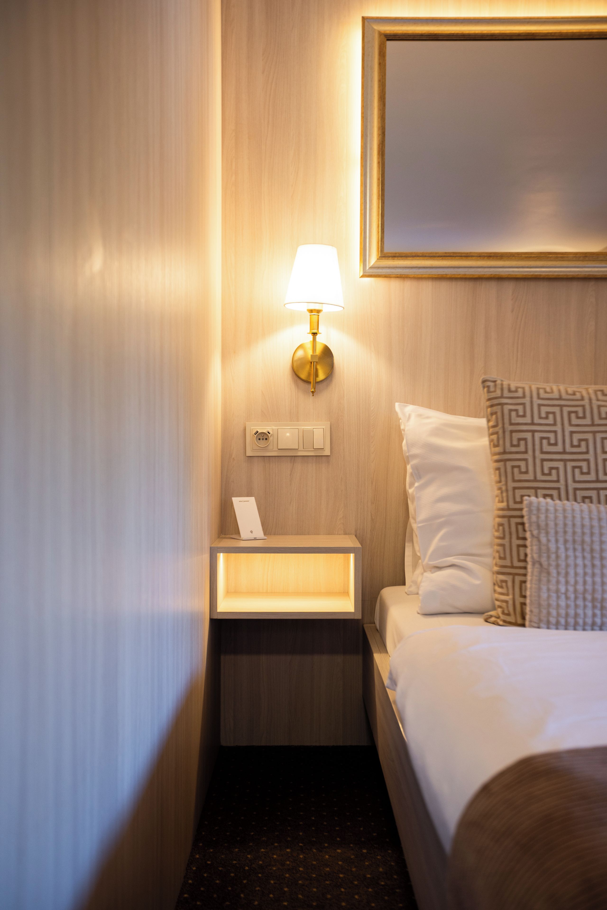 Habitación de hotel con cama, mesita de noche con lámpara, enchufe y panel de control en la pared.