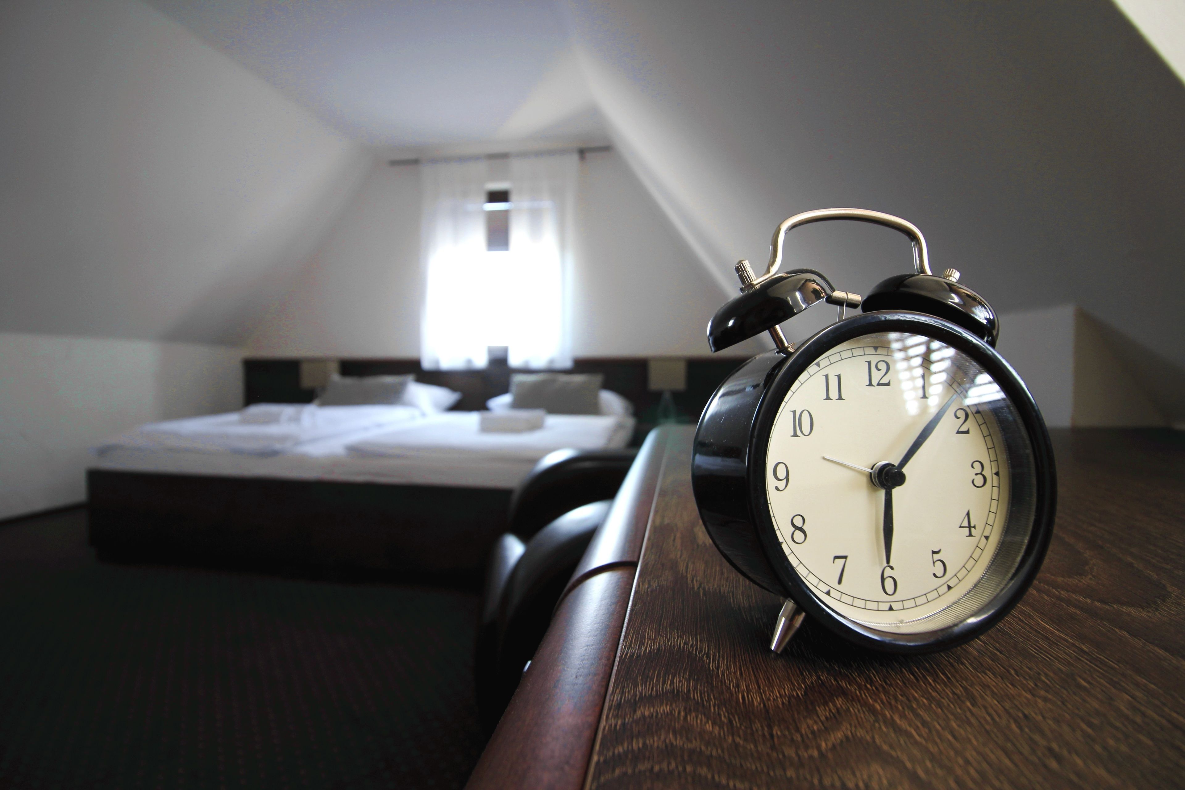 Una habitación con una cama y un despertador sobre una mesita de noche de madera.
