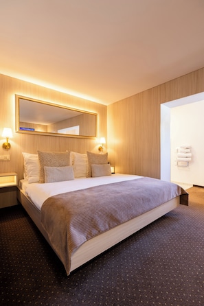 Quarto de hotel com uma cama grande, uma mesa de cabeceira com abajur, um espelho fixado na parede e uma porta para um banheiro.