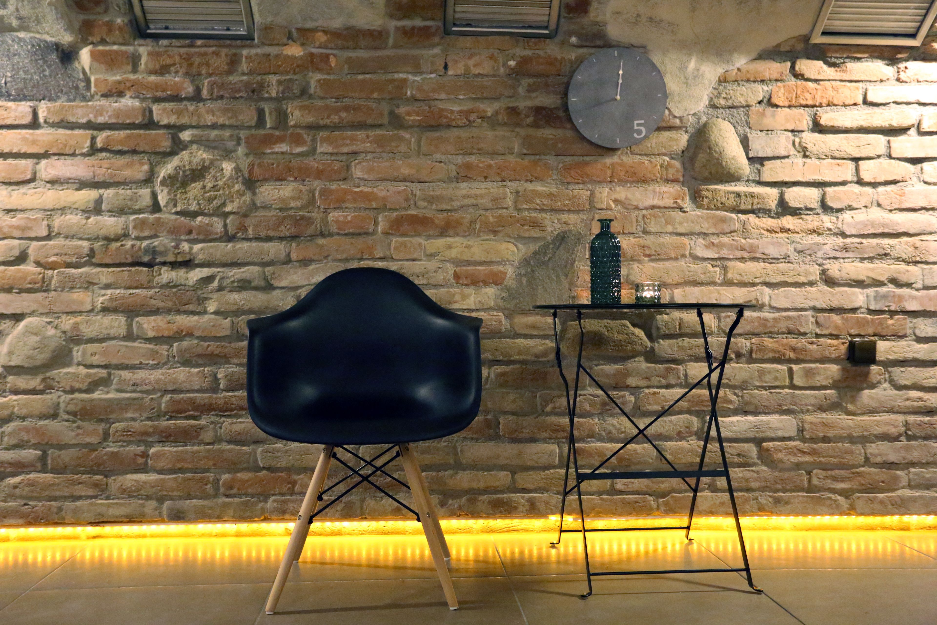 A room with a brick wall, a black chair, a small table with a bottle, and a clock.