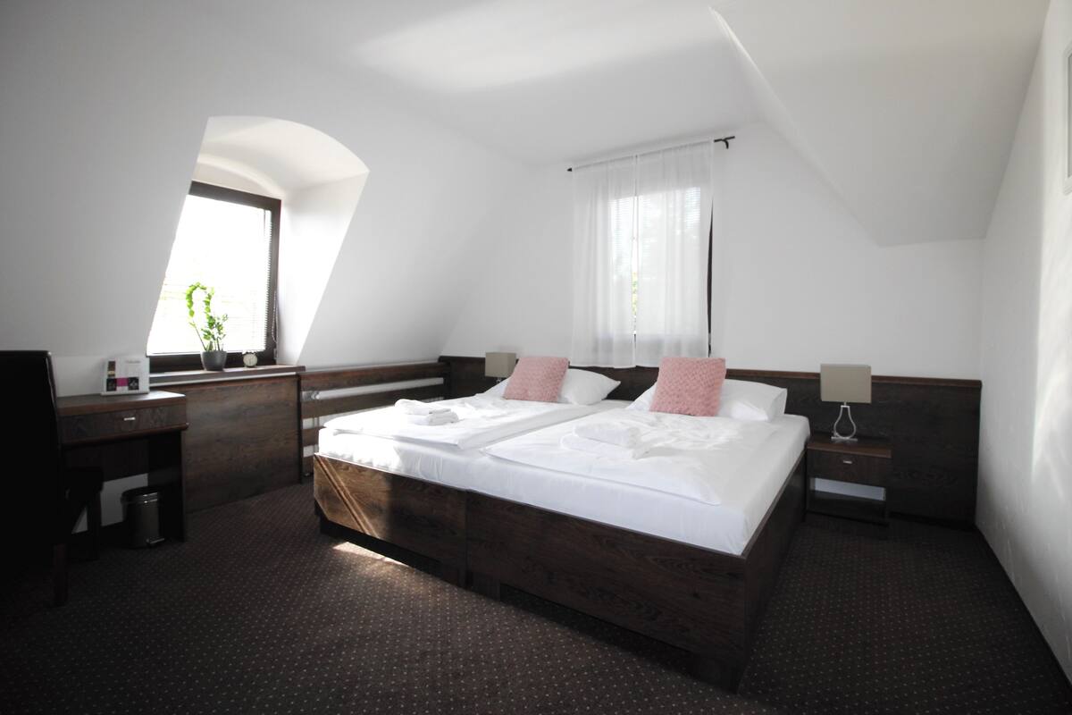 A hotel room with a large bed, a desk, a chair, a small table, a lamp, and a window with a plant.