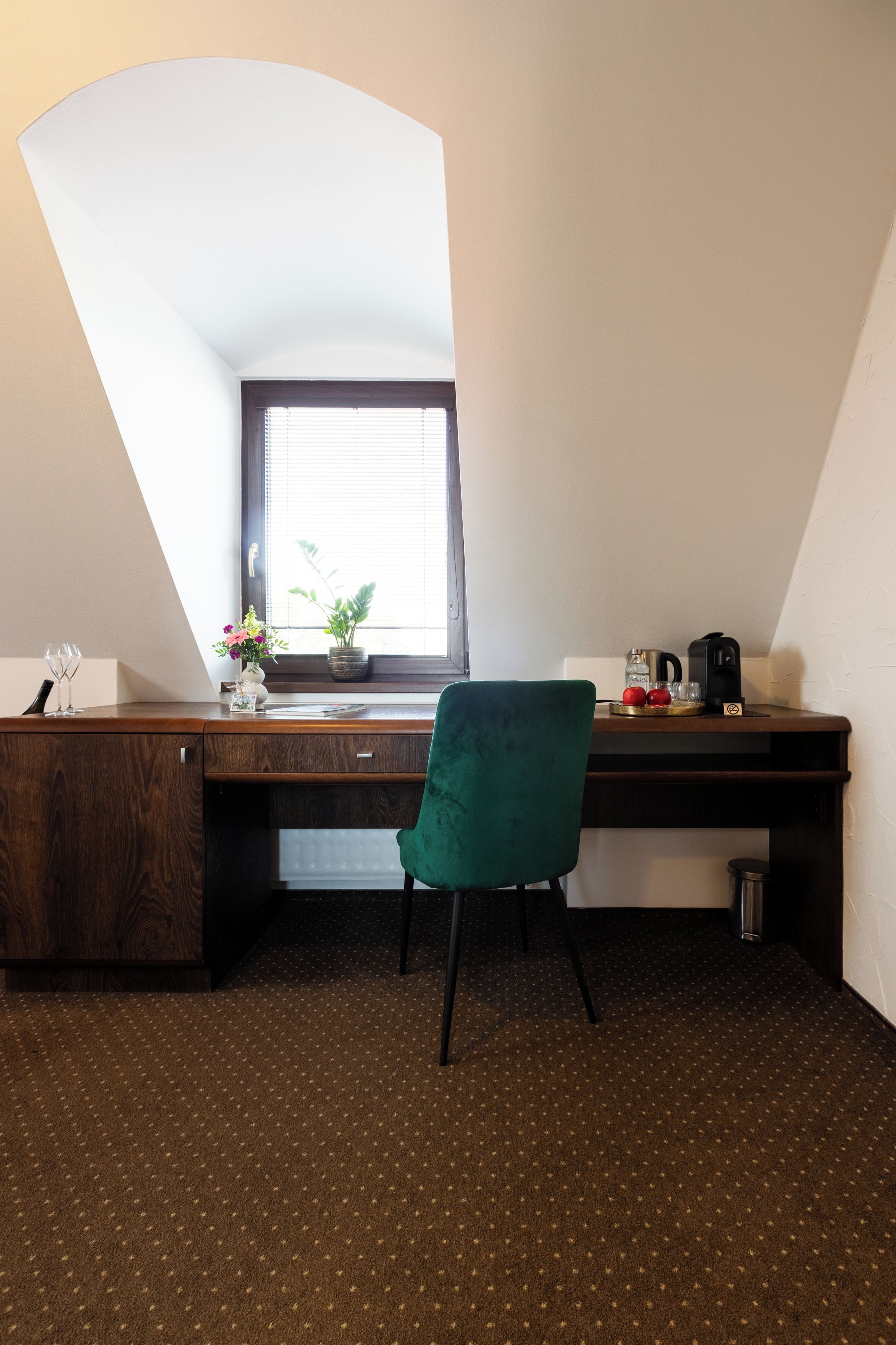 A room with a sloped ceiling, a desk with a computer, a chair, a window with blinds, and a small plant.