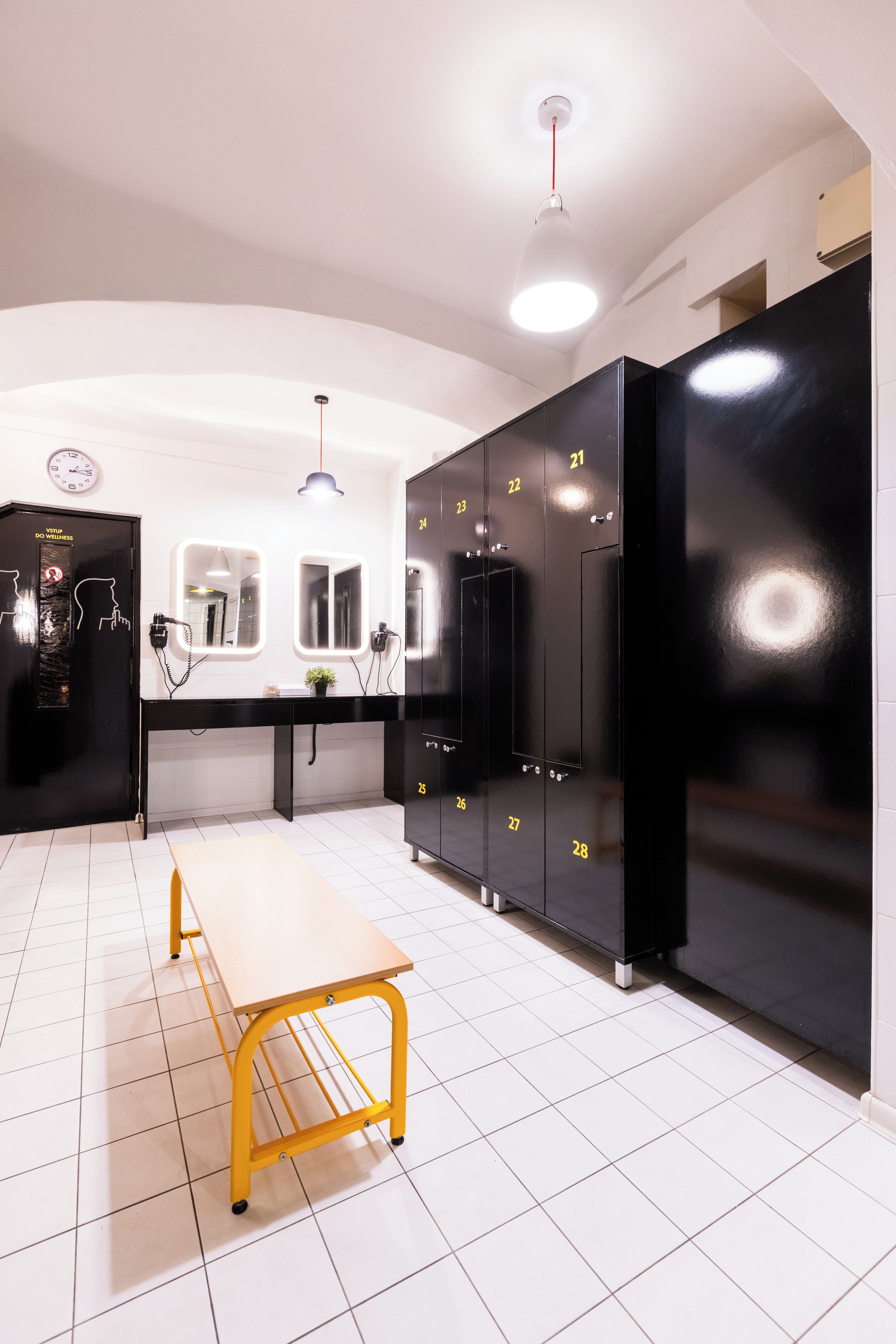 Un baño con piso de baldosas blancas, un banco de metal amarillo, taquillas negras numeradas del 21 al 28, un reloj y un espejo.