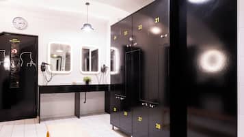 Un baño con piso de baldosas blancas, un banco de metal amarillo, taquillas negras numeradas del 21 al 28, un reloj y un espejo.