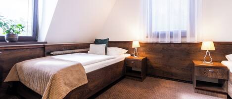 A hotel room with a bed, bedside tables, a lamp, a window with blinds, and a potted plant.