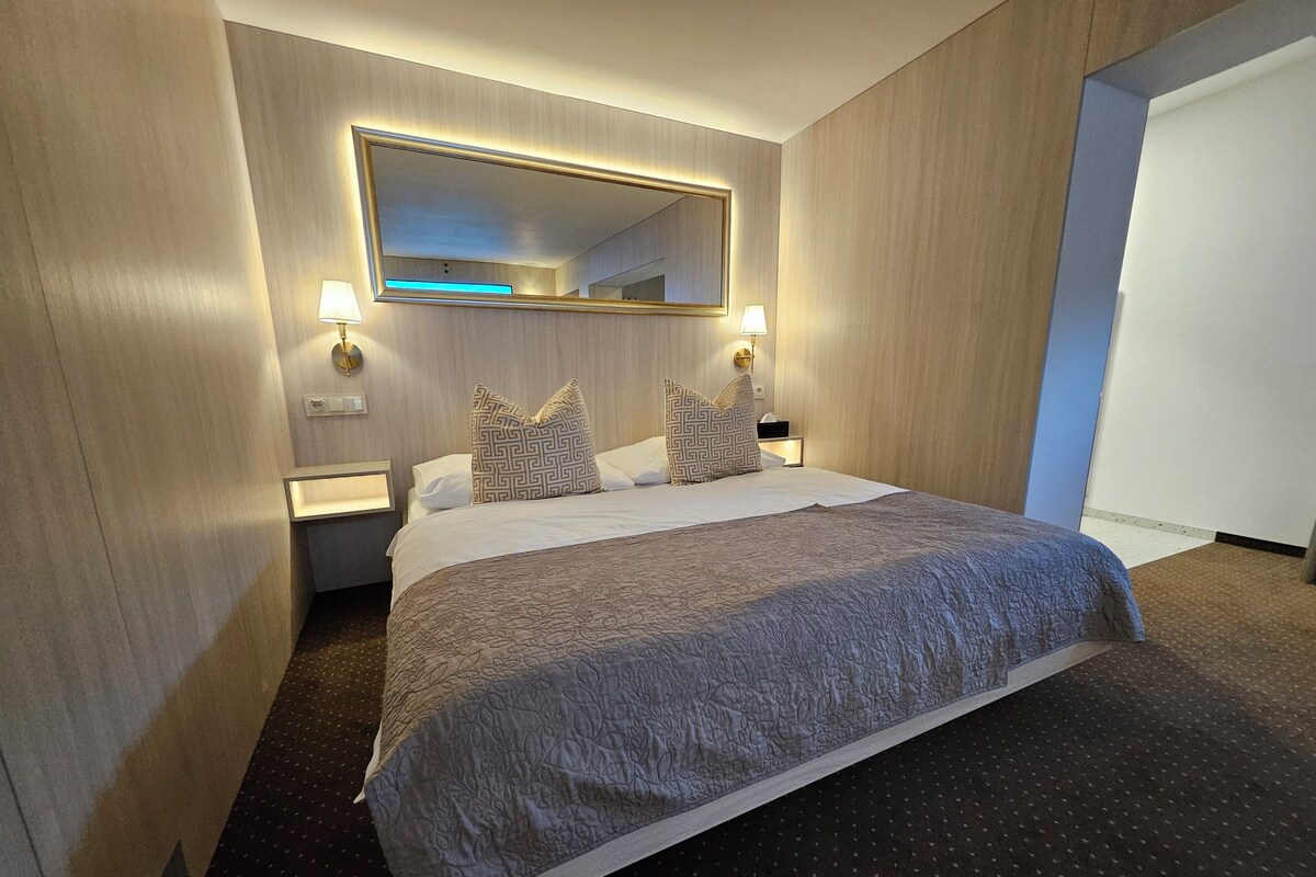 A hotel room with a large bed, a mirror above the bed, two bedside lamps, and a nightstand.