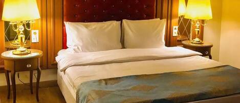 Una habitación de hotel con una cama grande, dos lámparas de noche, un cabecero tapizado en rojo y suelo de madera.