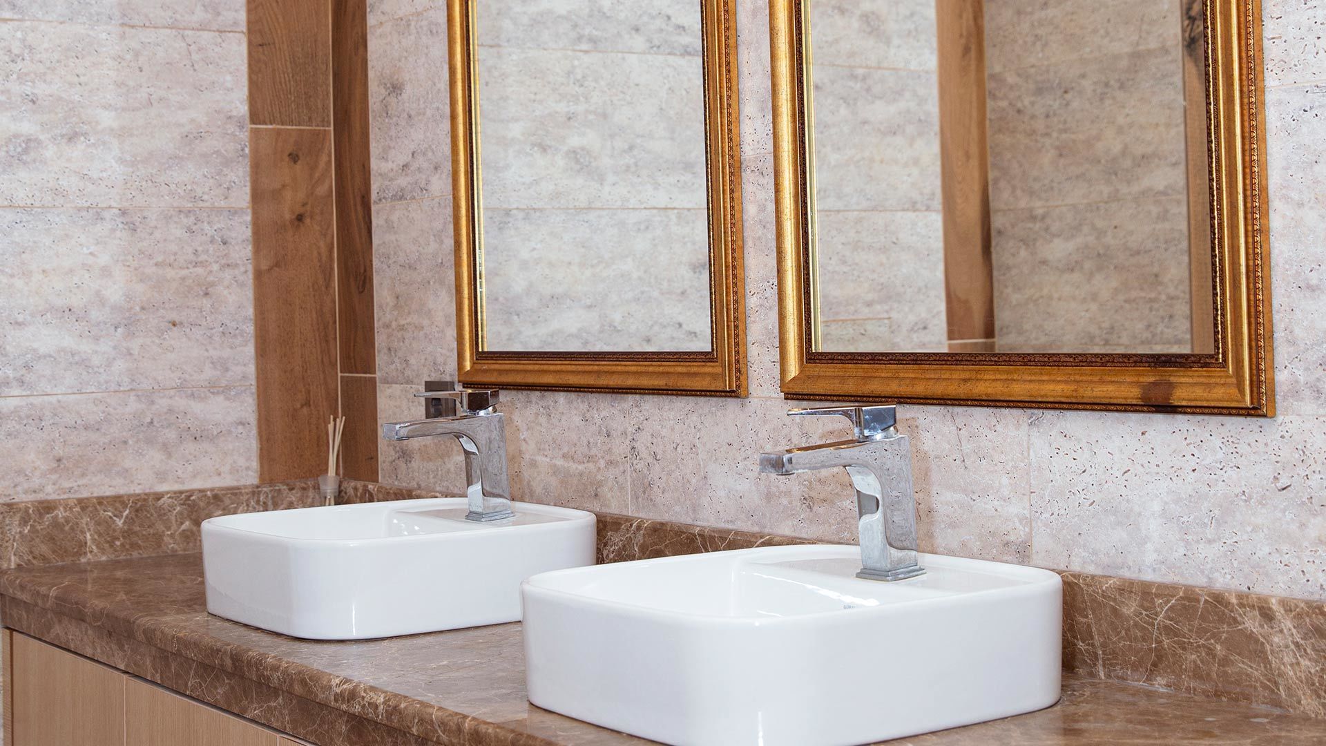 Un baño con dos lavamanos blancos, un espejo con marco dorado y encimera de mármol.