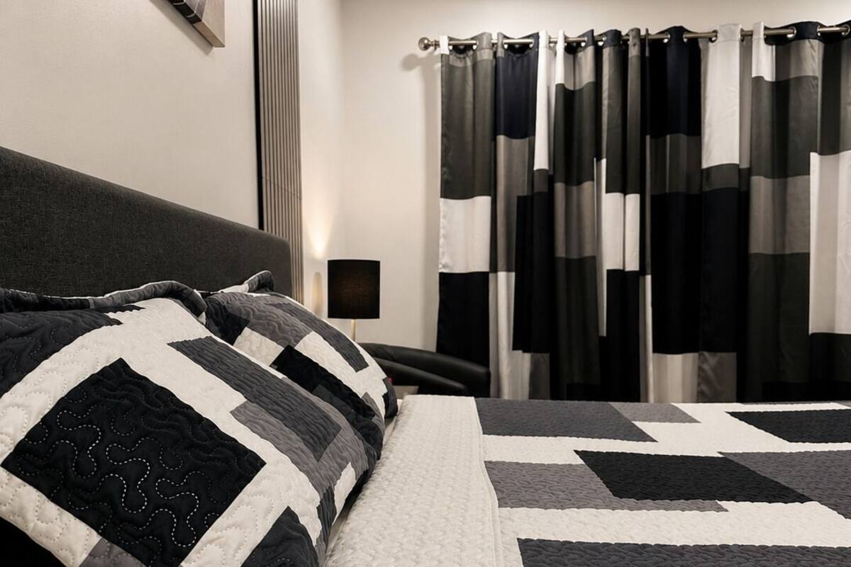Quarto com cama, mesa de cabeceira, luminária e janela com cortinas.