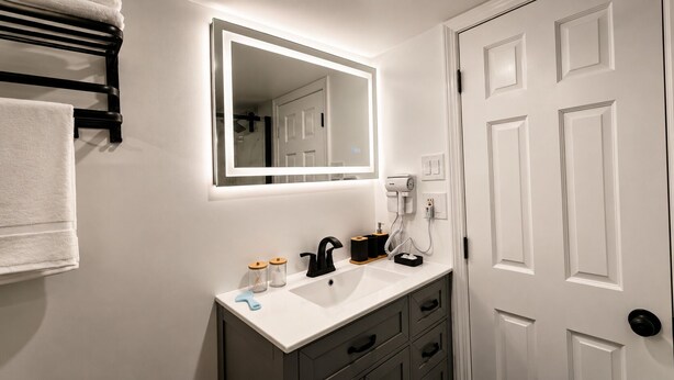A bathroom with a white sink, black faucet, and a mirror above it. There is a towel rack on the wall and a door to the right. - Lake George Residence LLC (South Glens Falls)