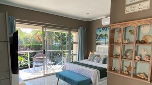 Une chambre avec un lit, un banc, une télévision, une porte-fenêtre coulissante donnant sur un balcon et un mur orné d’une étagère remplie de coquillages.