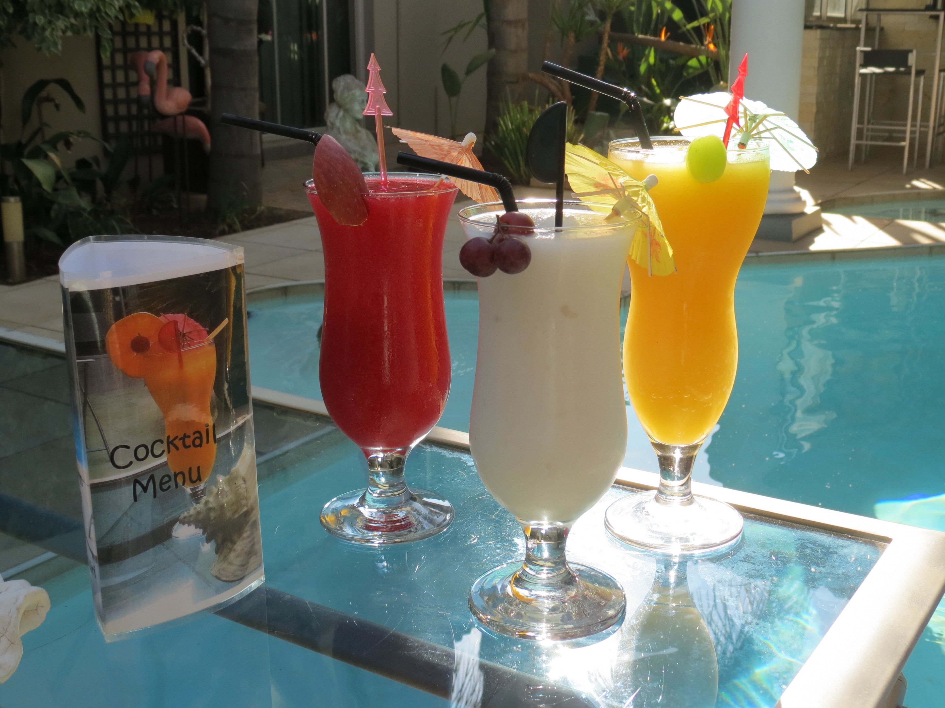 Three cocktails on a glass table by a poolside, with a cocktail menu visible.