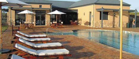 A pool area with lounge chairs and umbrellas, a brick patio, and a building with a grey roof.