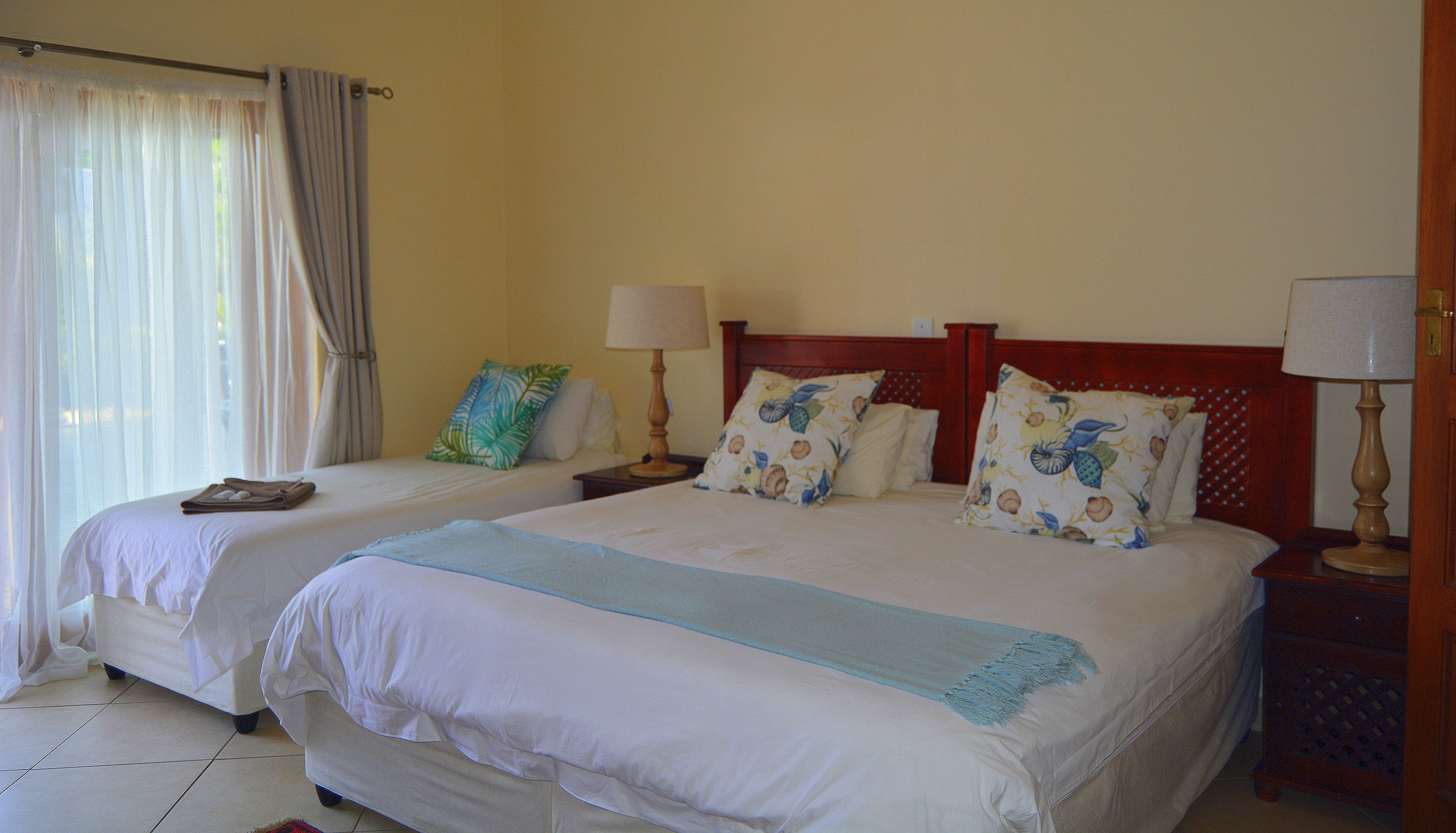 Habitación de hotel con dos camas, cada una con cabecero de madera, ropa de cama blanca y almohadas decorativas. Se ven mesitas de noche con lámparas y una ventana con cortinas.