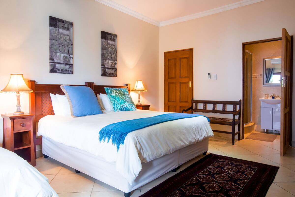 Quarto de hotel com uma cama grande, duas mesas de cabeceira com luminárias, um banco de madeira, um banheiro com pia e duas obras de arte penduradas na parede.