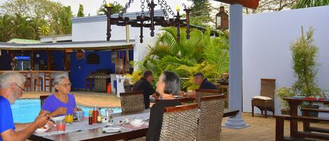 A dining area with a pool in the background, outdoor seating, and a hanging light fixture.