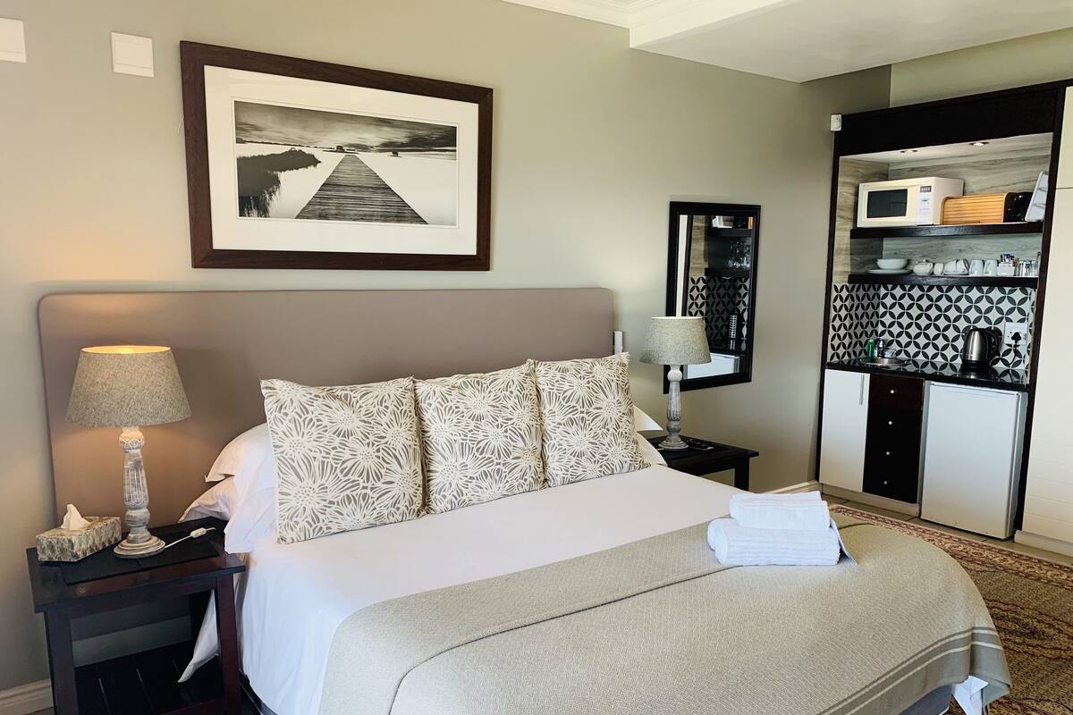 A hotel room with a bed, a nightstand, a lamp, a framed picture on the wall, a small table with a mirror, and a kitchenette area with a microwave, kettle, and cabinets.