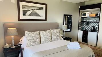 A hotel room with a bed, a nightstand, a lamp, a framed picture on the wall, a small table with a mirror, and a kitchenette area with a microwave, kettle, and cabinets.