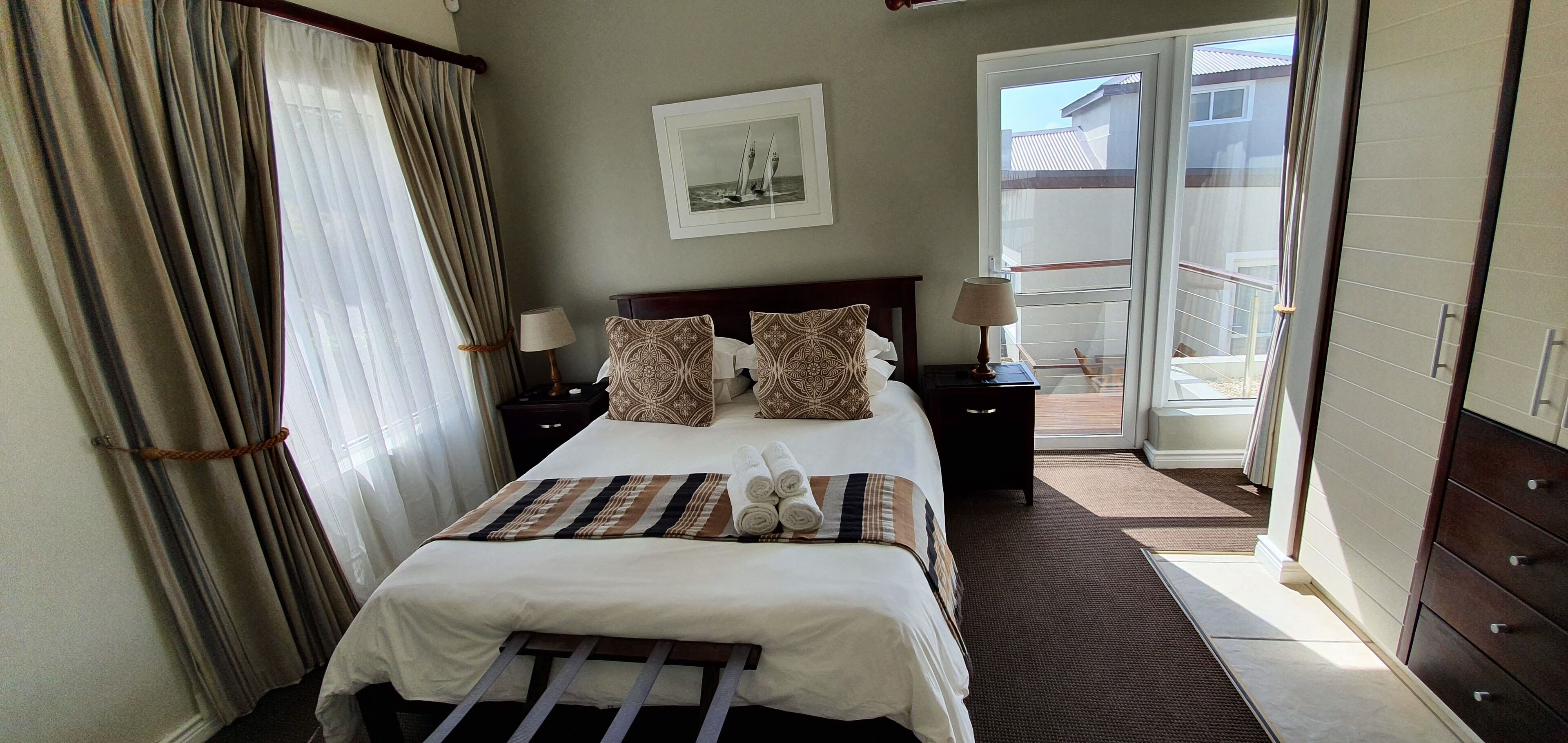 A hotel room with a bed, two bedside tables, a window with curtains, a door to a balcony, and a framed picture on the wall.