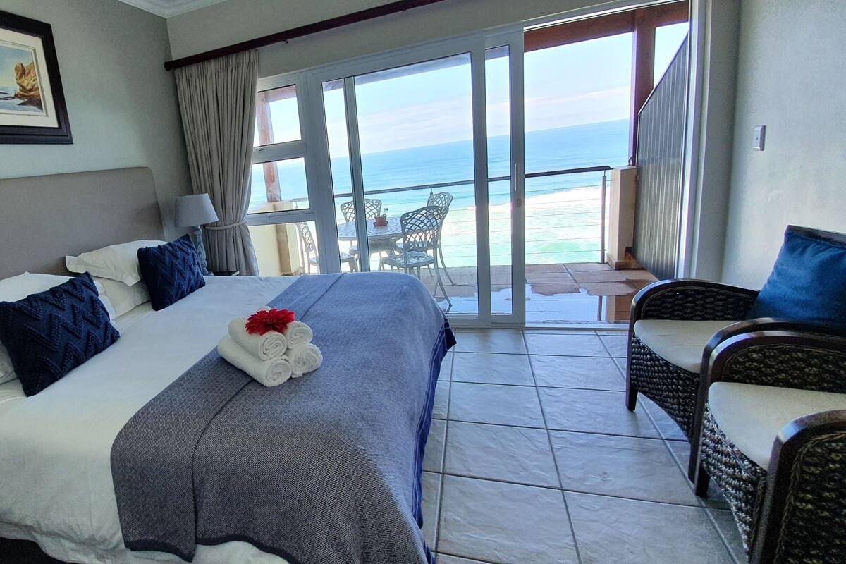 A hotel room with a bed, a chair, a table, and a view of the ocean.