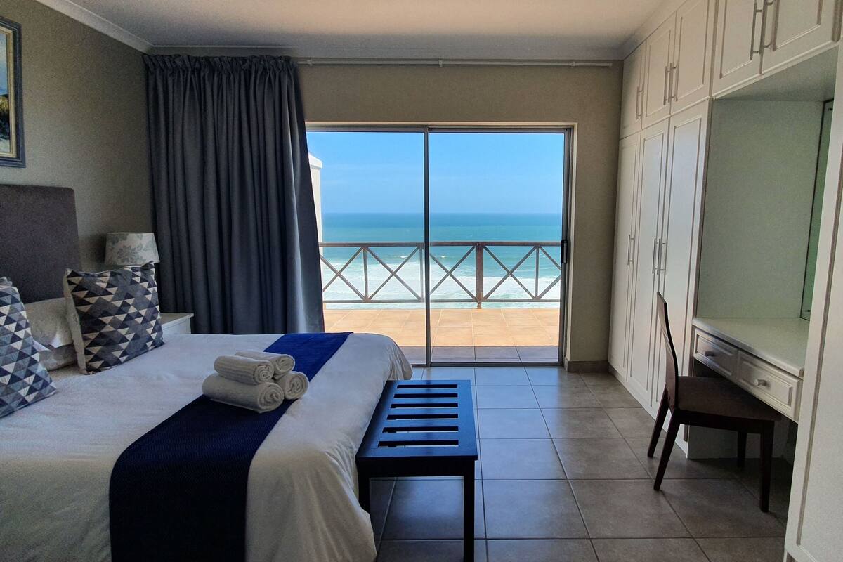 A hotel room with a bed, a desk, a TV, and a balcony with a view of the sea.