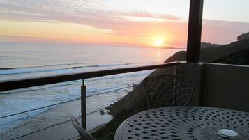 A balcony with a view of the ocean at sunset, a round metal table, and a chair.