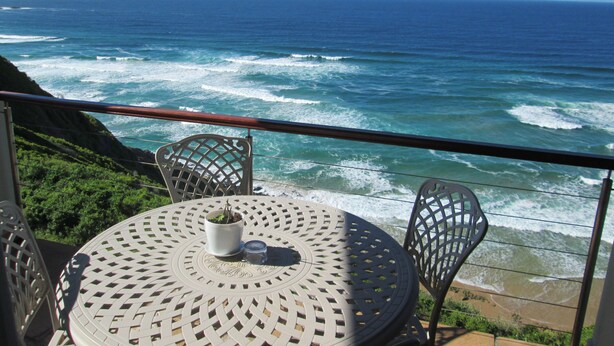 Une table ronde en métal, entourée de chaises, un pot de fleurs et un verre, le tout sur un balcon donnant sur l’océan.