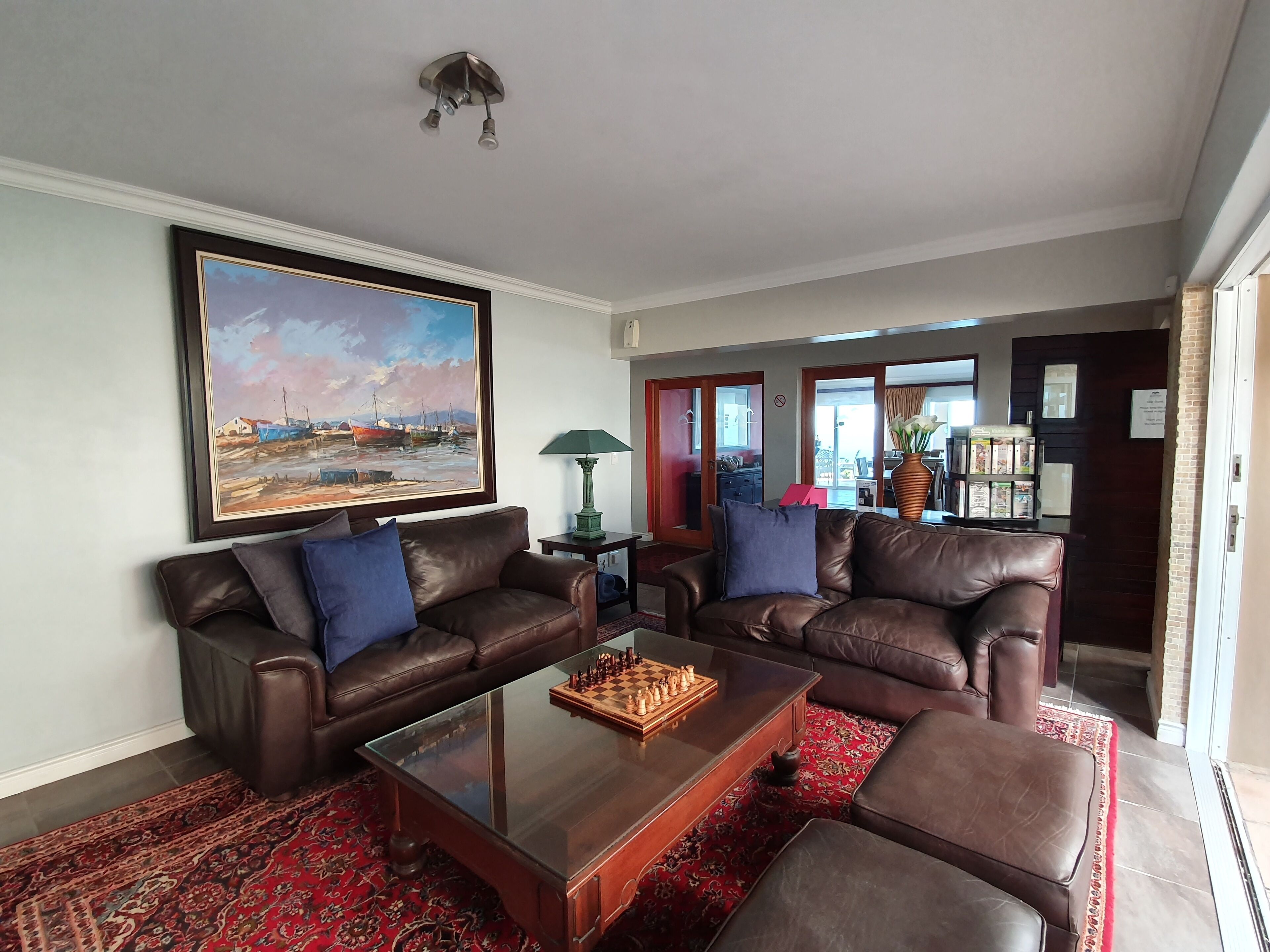 A living room with two brown leather sofas, a glass coffee table, a chessboard, a framed painting, a lamp, and a red patterned rug.