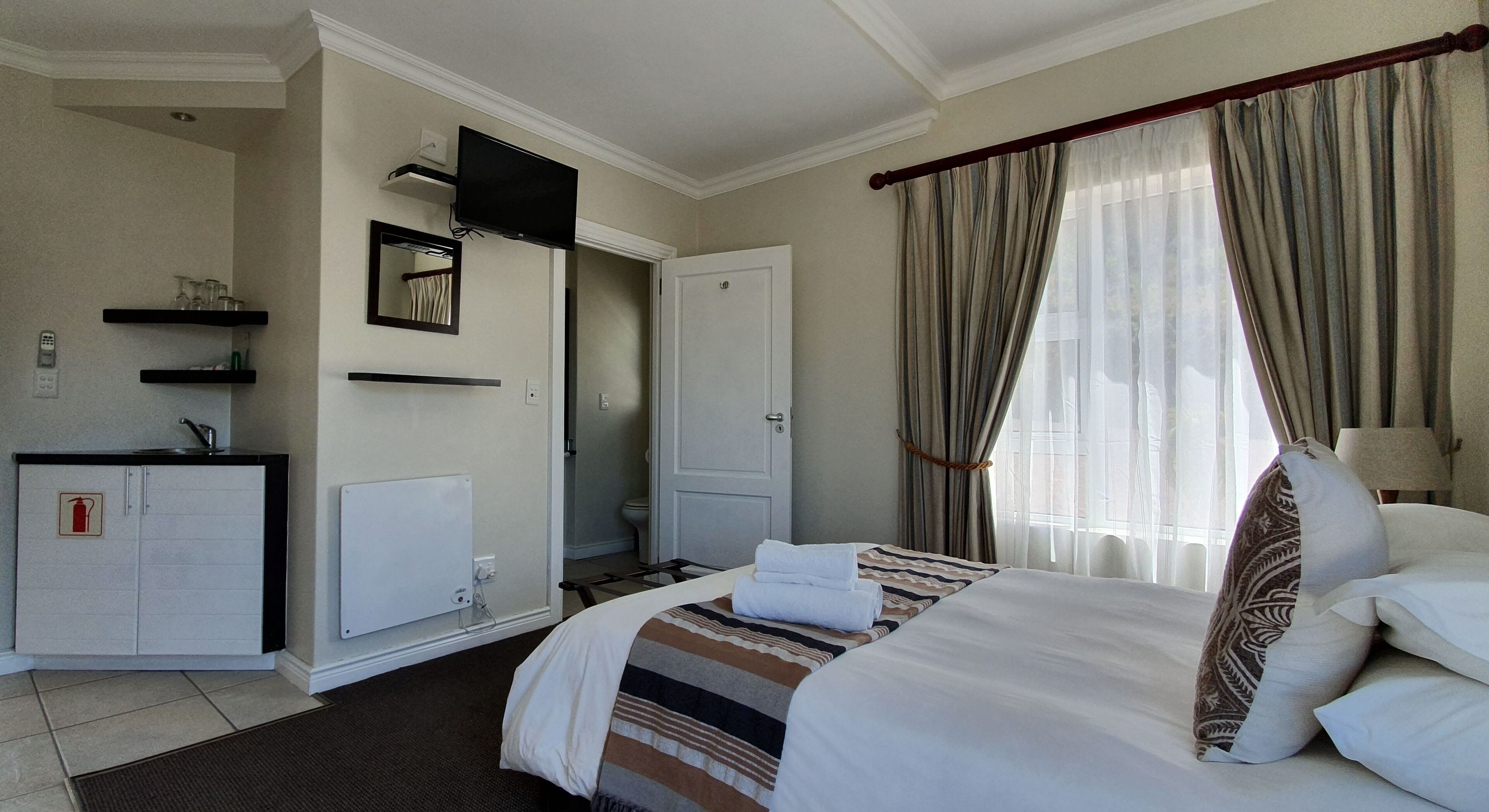 A hotel room with a bed, a desk with a computer, a mirror, a television, a radiator, and a window with curtains.