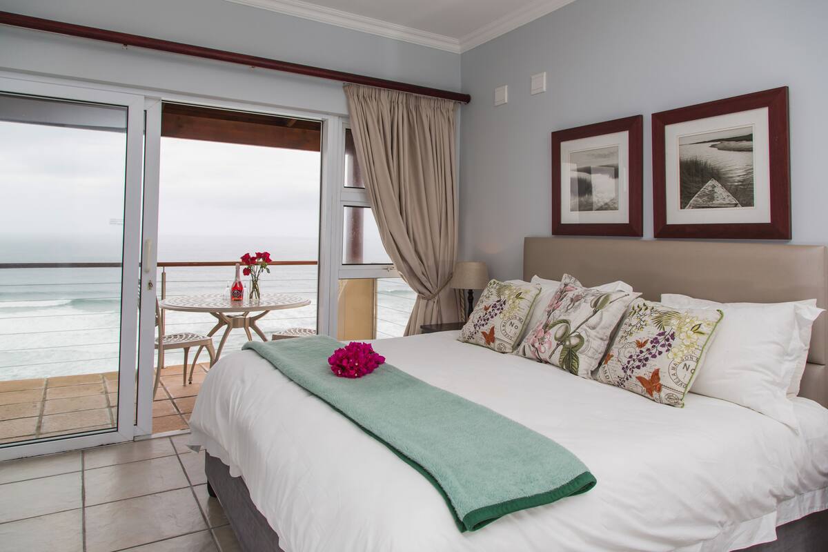 A hotel room with a bed, a balcony with a table and chairs, and two framed pictures on the wall.