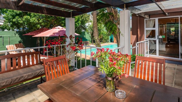 A wooden table with a vase of flowers, a pool, and a covered patio area with chairs and a table. - Brown Jug Guest House (Hermanus)