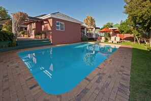 A swimming pool with a brick border, a red building, and a covered seating area with umbrellas. - Brown Jug Guest House (Hermanus)