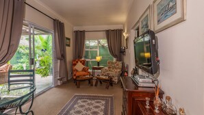 A cozy living room with a large TV, comfortable seating, and a view of greenery outside. - Brown Jug Guest House (Hermanus)